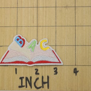 K&ouml;nnte beinhalten: Ein gestickter B&uuml;geleisen-Aufn&auml;her mit einem roten Buch mit den Buchstaben B, A und C in verschiedenen Farben. Das Buch ist ge&ouml;ffnet und die Buchstaben schauen aus den Seiten heraus.