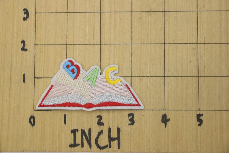 K&ouml;nnte beinhalten: Ein gestickter B&uuml;geleisen-Aufn&auml;her mit einem roten Buch mit den Buchstaben B, A und C in verschiedenen Farben. Das Buch ist ge&ouml;ffnet und die Buchstaben schauen aus den Seiten heraus.