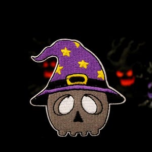 May include: A purple and yellow embroidered patch of a skull wearing a witch's hat. The skull has white eyes and a black nose.