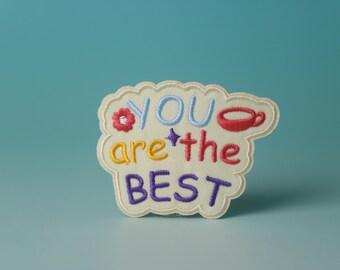 May include: A white felt patch with colorful embroidered text that reads "YOU are the BEST". The patch has a red flower, a blue star, and a red coffee cup.