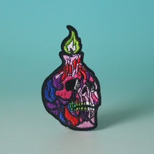 Puede incluir: Un parche bordado colorido que presenta una calavera con una vela en la parte superior. La calavera es rosa, morada, azul y roja, y la vela es verde y blanca.