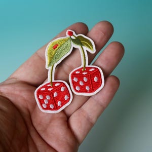 Puede incluir: Dos cerezas rojas en forma de dados con puntos blancos y tallos verdes. Las cerezas de dados est&aacute;n bordadas en una tela blanca.