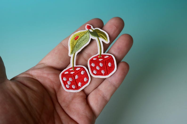 Puede incluir: Dos cerezas rojas en forma de dados con puntos blancos y tallos verdes. Las cerezas de dados est&aacute;n bordadas en una tela blanca.