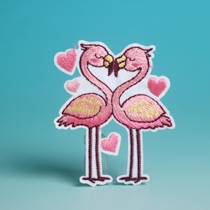 Pode incluir: Patch bordado com dois flamingos cor-de-rosa com asas amarelas, cujos pescoços formam um coração. Corações cor-de-rosa rodeiam os flamingos. O patch tem uma borda branca e está sobre um fundo azul-petróleo.