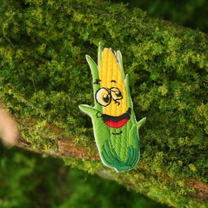 Pode incluir: Etiqueta bordada de uma espiga de milho de desenho animado com um rosto sorridente, folhas verdes e grãos amarelos. A etiqueta está sobre um fundo verde de musgo.