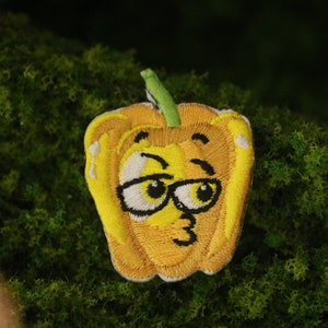 Puede incluir: Un parche bordado amarillo de un pimiento cartoon con un ojo guiñando y usando gafas. El pimiento tiene un tallo verde y un contorno marrón claro.