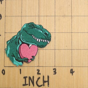 Pode incluir: Um dinossauro verde segurando um cora&ccedil;&atilde;o rosa. O dinossauro est&aacute; sorrindo e tem dentes brancos. O cora&ccedil;&atilde;o &eacute; uma forma simples sem detalhes.