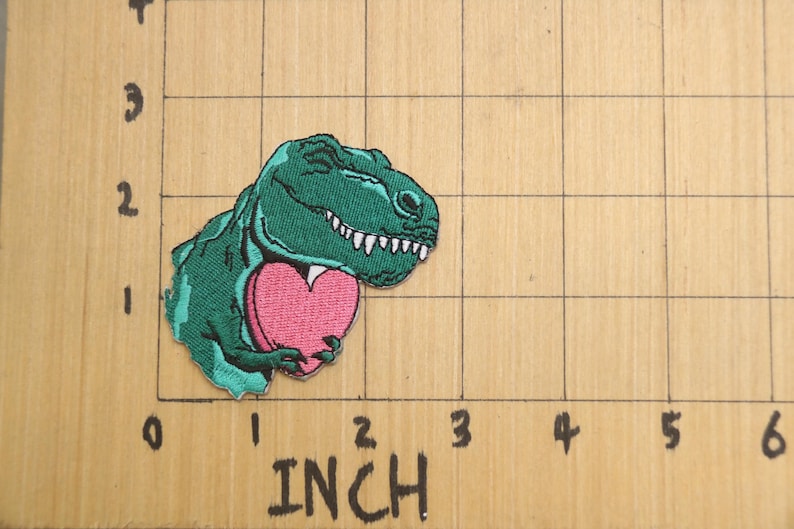 Pode incluir: Um dinossauro verde segurando um cora&ccedil;&atilde;o rosa. O dinossauro est&aacute; sorrindo e tem dentes brancos. O cora&ccedil;&atilde;o &eacute; uma forma simples sem detalhes.