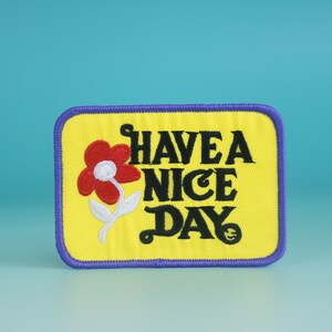 May include: Yellow embroidered patch with a purple border. The patch features a red flower with a white stem and the text "HAVE A NICE DAY".
