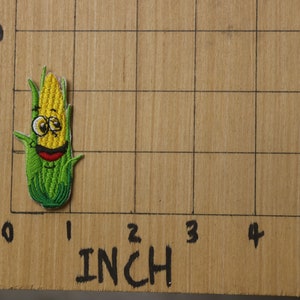 Peut inclure: Un patch brod&eacute; repr&eacute;sentant un &eacute;pi de ma&iuml;s avec un visage souriant, en jaune et vert. Le patch mesure environ 2,5 cm de haut et est pos&eacute; sur une surface en bois avec une grille.