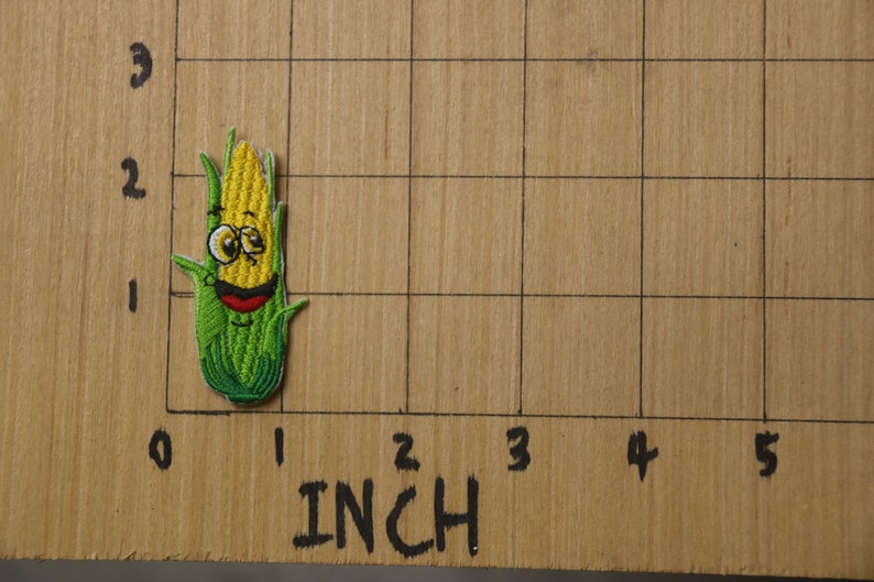 Peut inclure: Un patch brod&eacute; repr&eacute;sentant un &eacute;pi de ma&iuml;s avec un visage souriant, en jaune et vert. Le patch mesure environ 2,5 cm de haut et est pos&eacute; sur une surface en bois avec une grille.