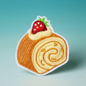以下が含まれることがあります： イチゴのショートケーキロールの刺繍パッチ。白、黄色、赤で刺繍されています。ロールは薄い茶色で、中心に白い渦巻きがあります。イチゴは赤で、白い種と緑の茎があります。