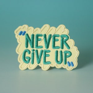 Può includere: Una toppa ricamata gialla con il testo "NEVER GIVE UP" in lettere verdi e virgolette blu.