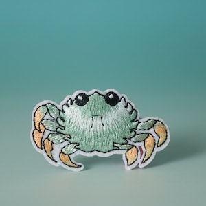 Op de afbeelding: Een groene geborduurde krab met oranje klauwen. De krab heeft een witte achtergrond en kijkt naar de kijker.