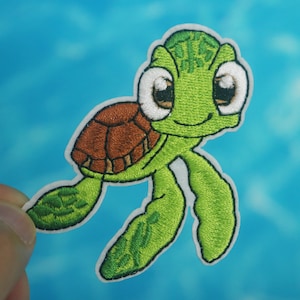 Può includere: Toppa ricamata verde e marrone da stirare con una tartaruga marina di cartone animato con occhi grandi e un sorriso.