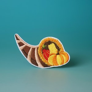 May include: Embroidered patch featuring a cornucopia design. The horn is a mix of brown and tan hues, with a white border. Inside, there's a pumpkin, a strawberry, and other harvest elements. Perfect for fall and Thanksgiving.