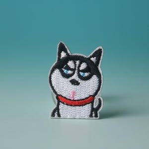May include: Embroidered patch of a cartoon husky dog with a grumpy expression. The patch has a white background with black and white details, blue eyes, and a red collar. The patch is approximately 5 cm tall.