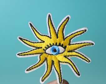 Op de afbeelding: Een gele geborduurde zon met een blauw oog in het midden. De zon heeft een witte omtrek en is omgeven door een blauwe achtergrond.