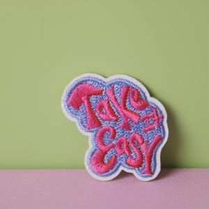 May include: A white embroidered patch with the words "Take it Easy" in pink lettering. The patch has a blue outline.