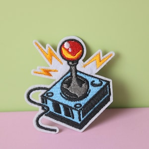 May include: An embroidered patch depicting a retro video game joystick in blue and black, with a red button and yellow lightning bolts.