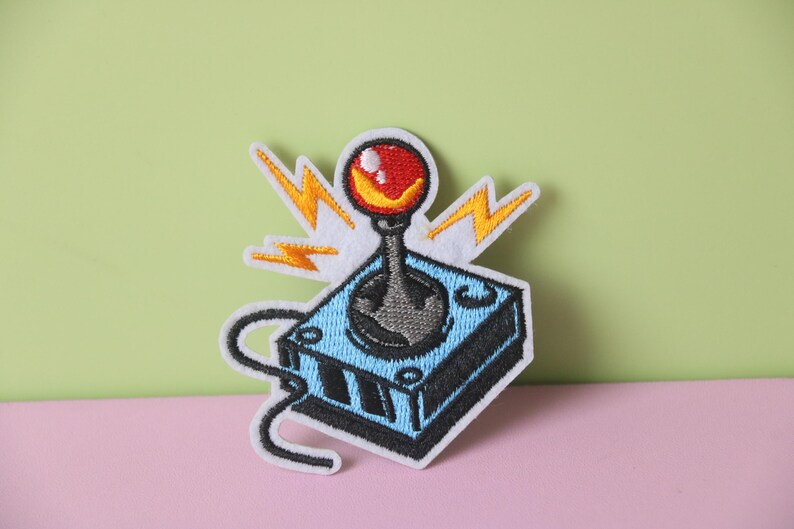 May include: An embroidered patch depicting a retro video game joystick in blue and black, with a red button and yellow lightning bolts.