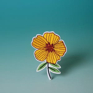 May include: A yellow embroidered flower with a brown centre and green leaves against a blue background.