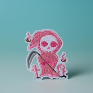 Puede incluir: Parche de hierro bordado de color rosa de un Grim Reaper de dibujos animados que sostiene una guadaña. El Grim Reaper lleva una túnica con capucha rosa y tiene una calavera blanca con ojos rosas.