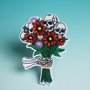 Op de afbeelding: Geborduurde patch met een boeket bloemen met schedelgezichten. De bloemen zijn rood met gele centra en paarse en roze accenten. Het boeket wordt vastgehouden door een skelet hand, met groene bladeren en stelen.