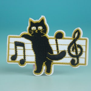May include: Black cat embroidered patch with a musical note and treble clef on a white background with gold stitching.