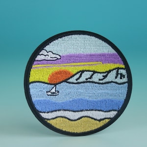 May include: A round embroidered patch with a sunset scene over the ocean. The patch features a yellow sun, purple and yellow sky, blue water, and a white sailboat. The patch is surrounded by a black border.