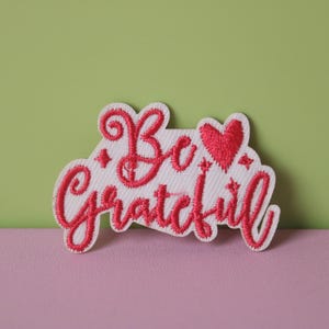 May include: White fabric patch with pink embroidered text that reads "Be grateful" with a pink heart in the middle.