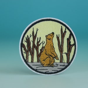 Puede incluir: Un parche redondo bordado con un oso marrón de pie en un bosque de ramas marrones. El fondo es de color amarillo claro.