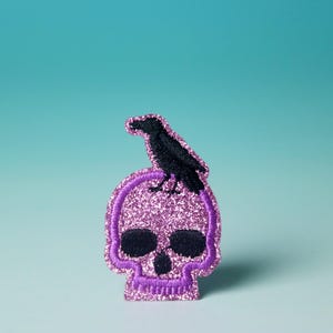 May include: A glittery purple skull patch with a black crow perched on top. The skull has black eye sockets and a purple outline. The patch is likely for clothing or accessories.