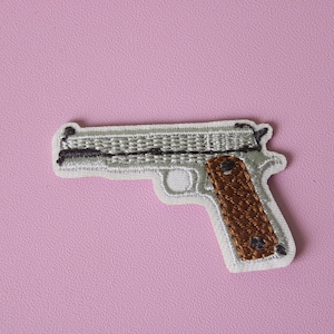 May include: A white embroidered patch of a silver handgun with a brown wooden handle.