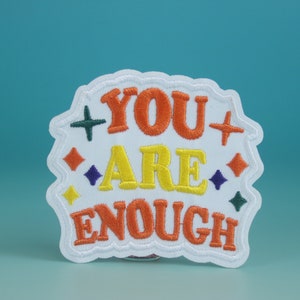 Pode incluir: Etiqueta branca bordada com o texto "YOU ARE ENOUGH" em letras laranja, amarelo e verde. A etiqueta é cercada por estrelas coloridas.