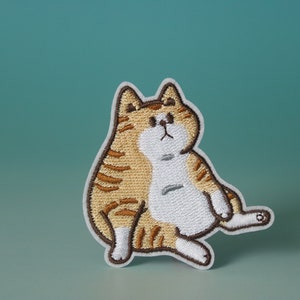 Może przedstawiać: Naszywka z haftem przedstawiająca kreskówkową kotkę w brązowo-białe paski. Kotka siedzi ze skrzyżowanymi łapkami i ma biały brzuszek.