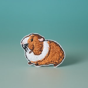 Pode incluir: Etiqueta bordada para passar a ferro de um porquinho-da-índia castanho e branco. O porquinho-da-índia está virado para a esquerda e tem uma mancha branca no peito.