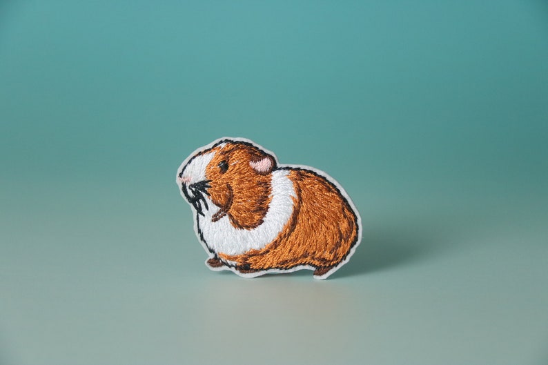 K&ouml;nnte beinhalten: Gestickter B&uuml;gelaufkleber mit einem braunen und wei&szlig;en Meerschweinchen. Das Meerschweinchen blickt nach links und hat einen wei&szlig;en Fleck auf der Brust.