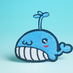 May include: Embroidered patch of a cartoon whale in blue, white, and black. The whale has a smiling face with rosy cheeks and a small spout. The patch is designed for clothing or accessories.