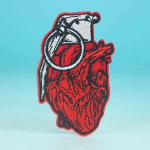 May include: Red embroidered patch of a heart shaped grenade with a white pull pin.
