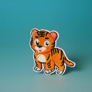 Op de afbeelding: Een geborduurde patch met een cartoon-tijger met oranje en zwarte strepen, witte poten en een roze neus. De tijger staat op een blauwe achtergrond.