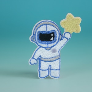 Może przedstawiać: Biała haftowana naszywka przedstawiająca astronautę w niebieskim hełmie trzymającego żółtą gwiazdę. Astronauta ma na sobie biały skafander kosmiczny z niebieskimi akcentami.