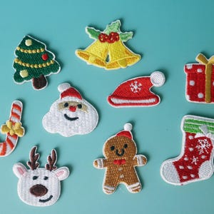 Könnte beinhalten: Eine Sammlung gestickter Weihnachtspatches. Sie zeigen einen Weihnachtsbaum, Glocken, einen Weihnachtsmann, ein Geschenk, eine Zuckerstange, ein Rentier, einen Lebkuchenmann und einen Strumpf. Die Patches sind in Rot, Grün, Weiß und Gold gehalten.