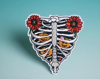 May include: Embroidered patch with a white rib cage design and black outlines. Two red sunflowers are on either side of the rib cage. Inside the rib cage are three orange jack-o'-lanterns. A small pink flower is also inside the rib cage.