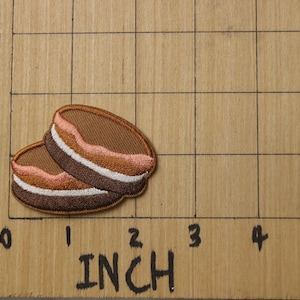 May include: Two brown and white embroidered patches shaped like a stack of pancakes. The patches are about 1 inch in diameter and are on a brown wooden surface with a grid.