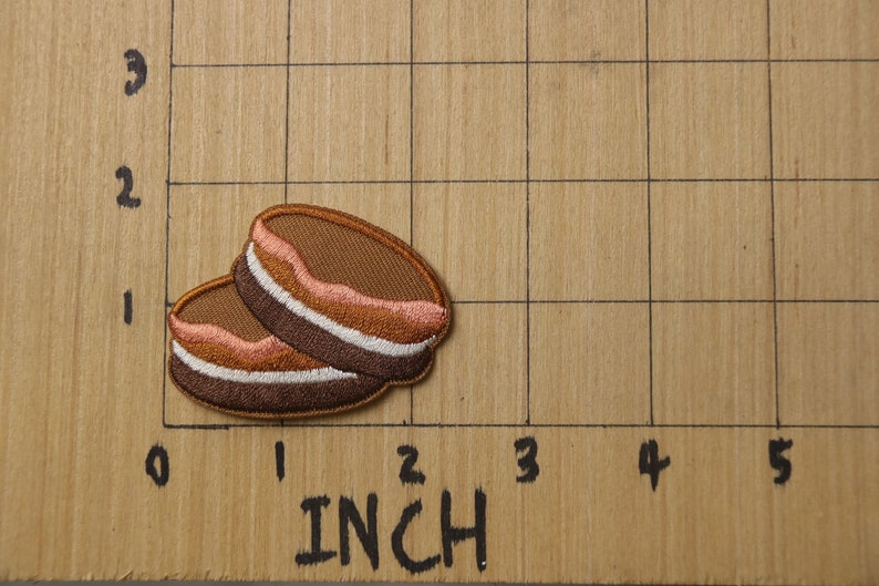 May include: Two brown and white embroidered patches shaped like a stack of pancakes. The patches are about 1 inch in diameter and are on a brown wooden surface with a grid.