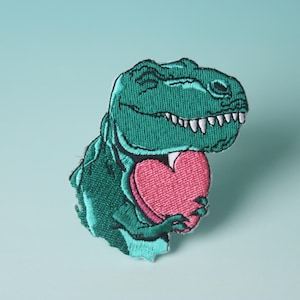 Könnte beinhalten: Ein grüner gestickter Dinosaurier hält ein pinkfarbenes Herz. Der Dinosaurier lächelt und hat weiße Zähne. Das Herz ist eine einfache Form ohne Details.