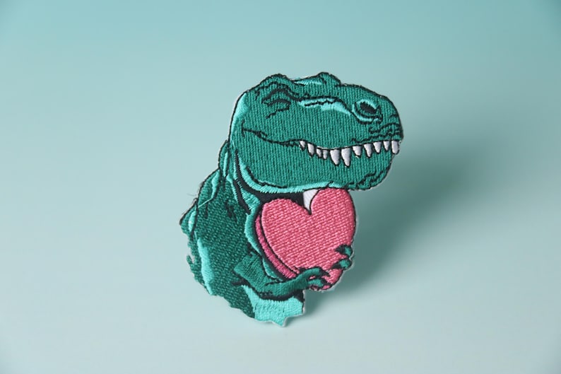 Pode incluir: Um dinossauro verde bordado segurando um cora&ccedil;&atilde;o rosa. O dinossauro est&aacute; sorrindo e tem dentes brancos. O cora&ccedil;&atilde;o &eacute; uma forma simples sem detalhes.