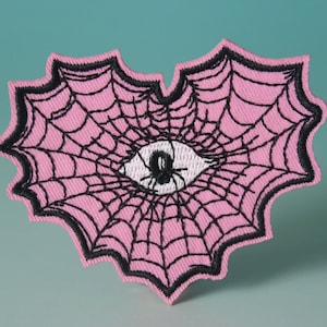 May include: Pink heart-shaped embroidered patch with a black spider web and a white eye in the center. The spider is black with a white body.