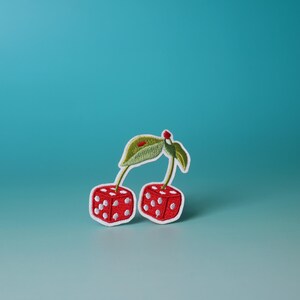 Puede incluir: Un parche bordado con dos cerezas, cada una con forma de dado, con puntos rojos y blancos. Los tallos y las hojas son verdes.